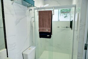 A modern bathroom with a glass shower enclosure, a toilet, and a towel rack with a brown towel. - Jubarte Chalés | Confort near the beach (Ilhabela)