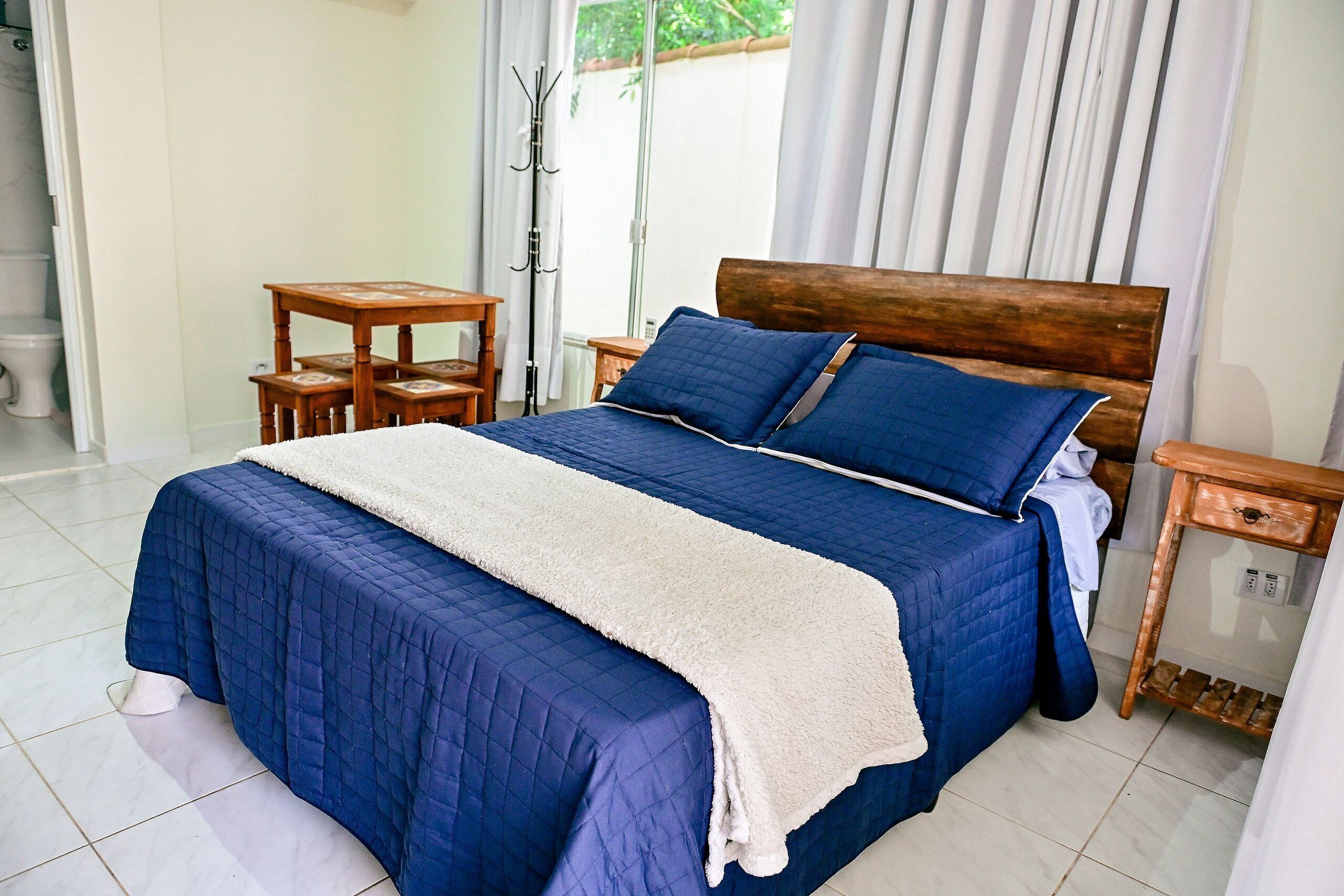Uma cama bem arrumada com um edredom azul e um xale branco, uma mesinha de cabeceira de madeira com uma luminária, e uma pequena mesa com uma cadeira.