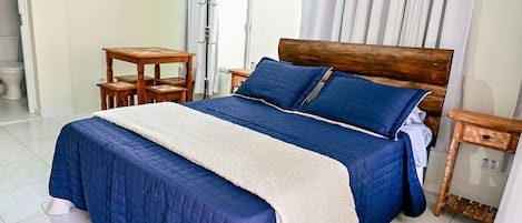 A neatly made bed with a blue quilt and a white throw, a wooden nightstand with a lamp, and a small table with a chair.