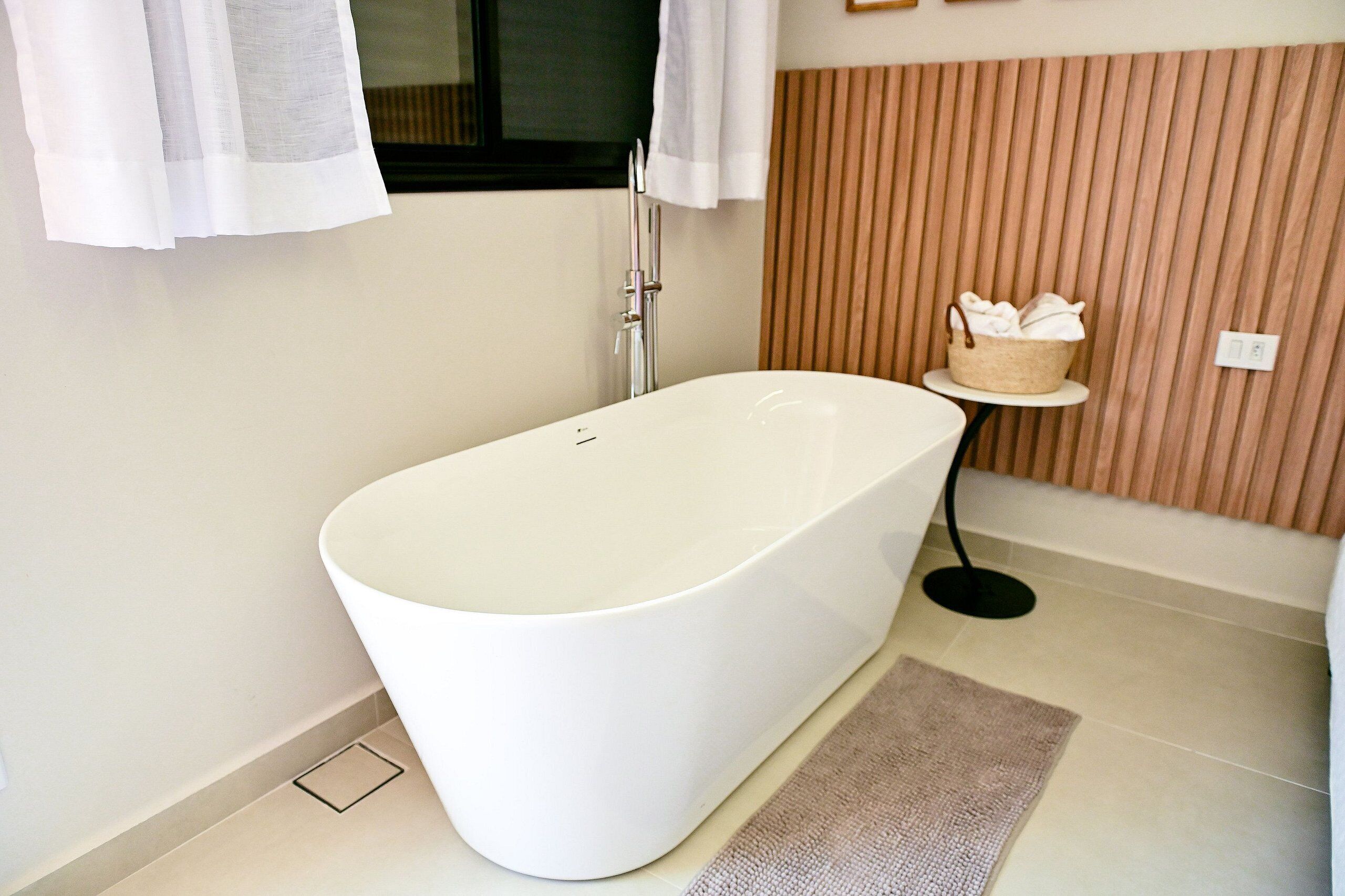 Un baño moderno con una bañera independiente, un perchero, una mesita con una cesta y una ventana con cortinas.