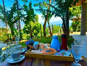 A breakfast tray with food and drinks on a wooden table outdoors with a view of the sea and greenery.