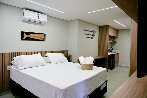 A hotel room with a bed, a wall-mounted air conditioner, a wooden headboard, a wall-mounted towel rack, a small desk with a chair, and a woven basket with towels.