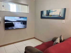 A living room with a flat-screen TV mounted on the wall, a couch with pillows, and a painting above the TV. - 403 Apto Versátil e Aconchegante (Belém)