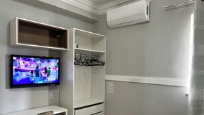 A room with a TV mounted on the wall, a desk with a chair, a wardrobe with hanging space, and an air conditioning unit. - 403 Apto Versátil e Aconchegante (Belém)