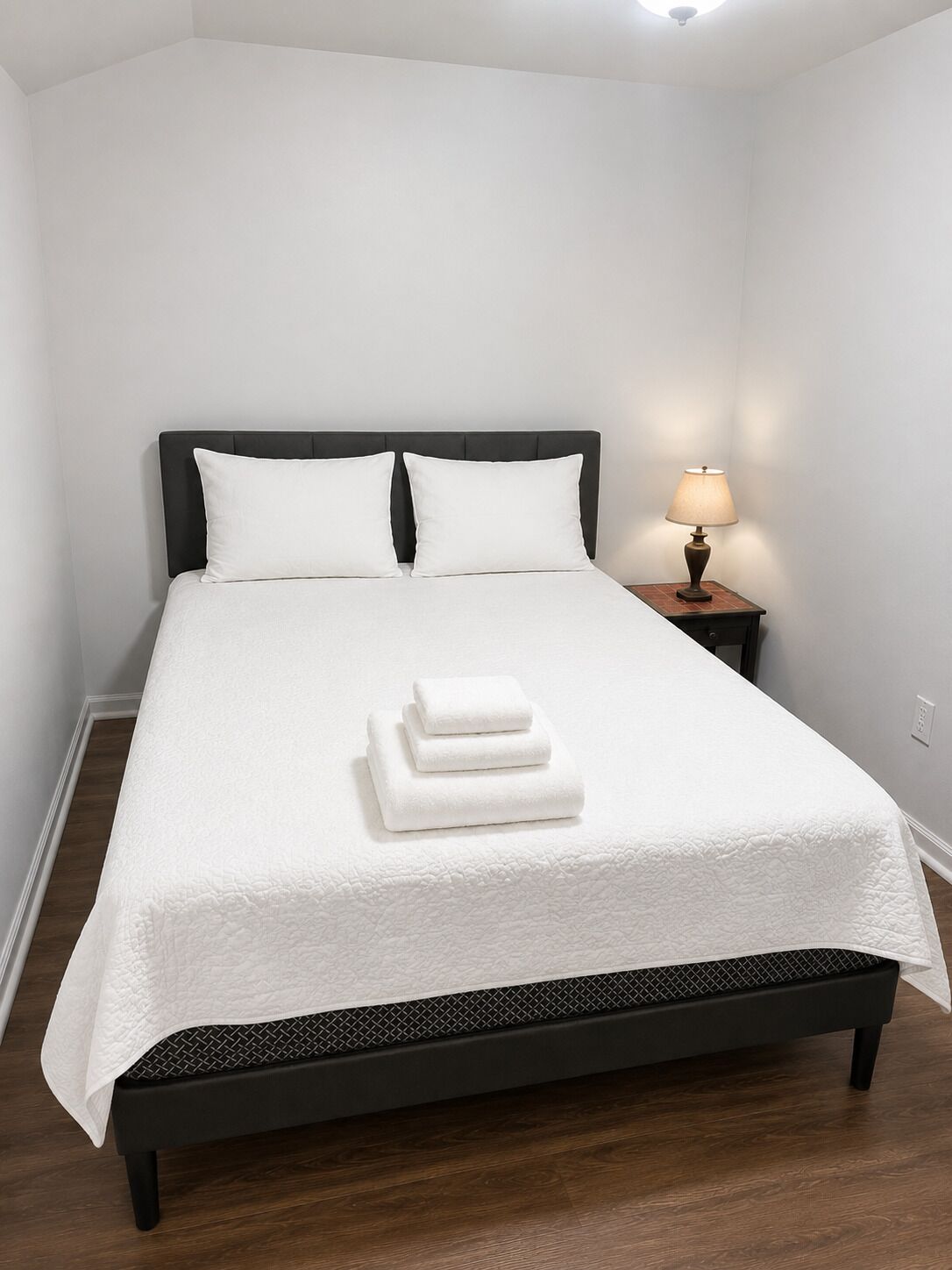 A neatly made bed with a white bedspread and pillows, a bedside lamp, and a stack of folded towels.