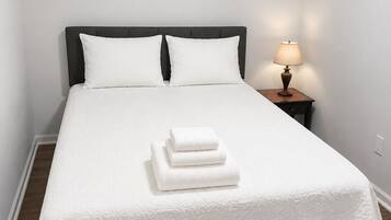 A neatly made bed with a white bedspread and pillows, a bedside lamp, and a stack of folded towels.