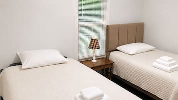 A hotel room with two beds, a bedside table with a lamp, and a window with blinds.