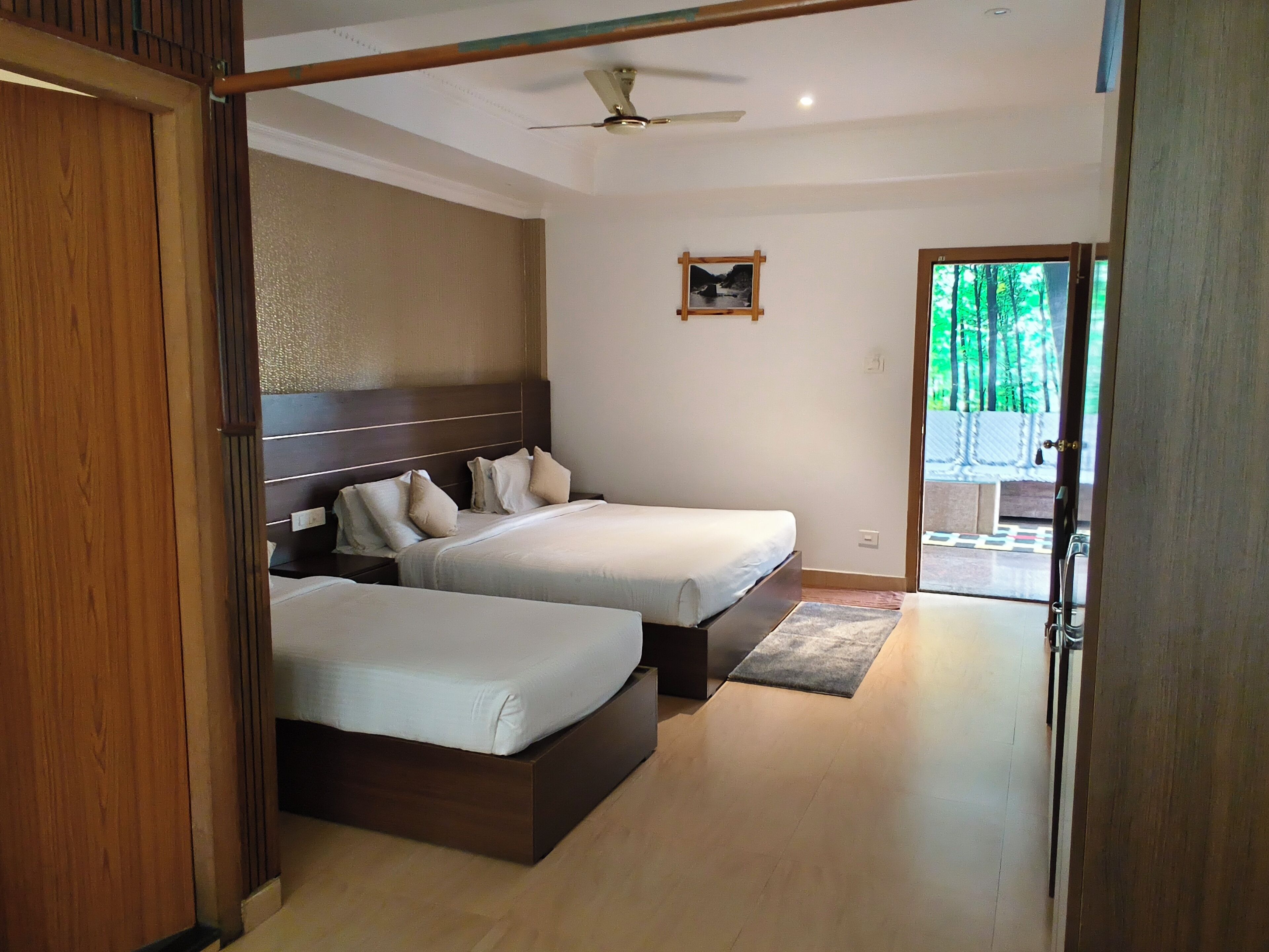 A hotel room with two beds, a ceiling fan, a small picture on the wall, and a view of greenery outside.