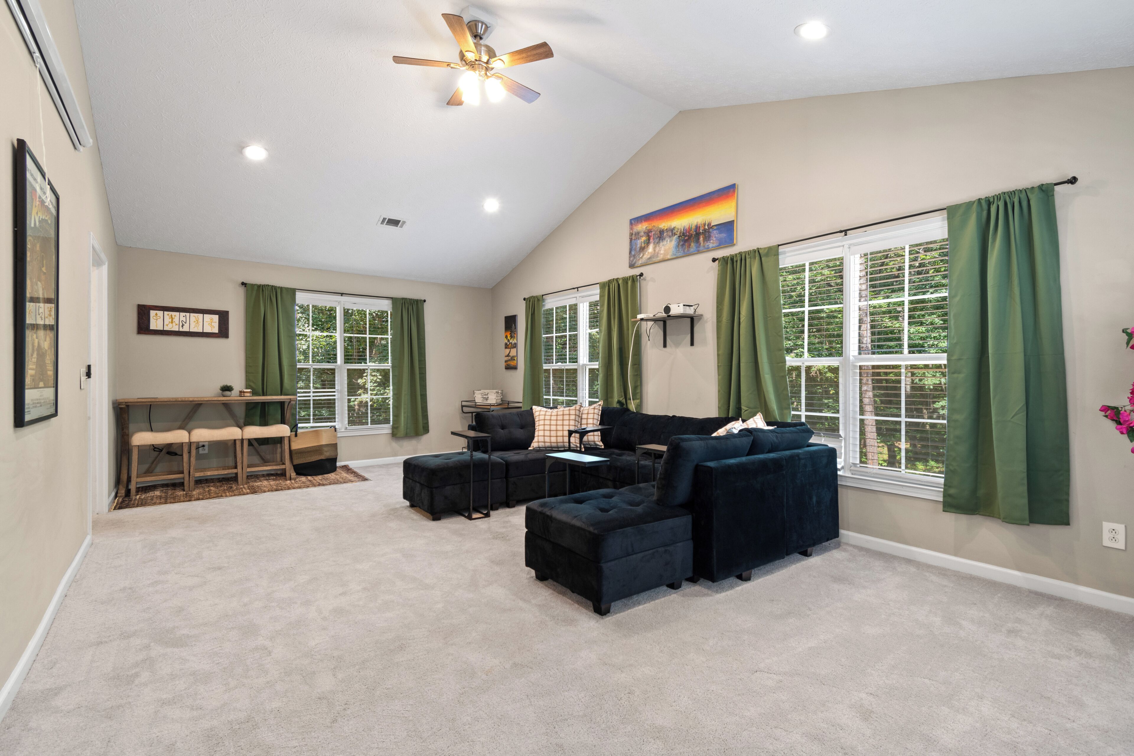 Un salon spacieux doté d’un ventilateur de plafond, de grandes fenêtres avec des rideaux verts, d’un canapé d’angle noir et d’une petite table avec une chaise.
