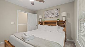 A bedroom with a bed, a ceiling fan, a door, a wall-mounted shelf with decorative items, and a window with blinds.