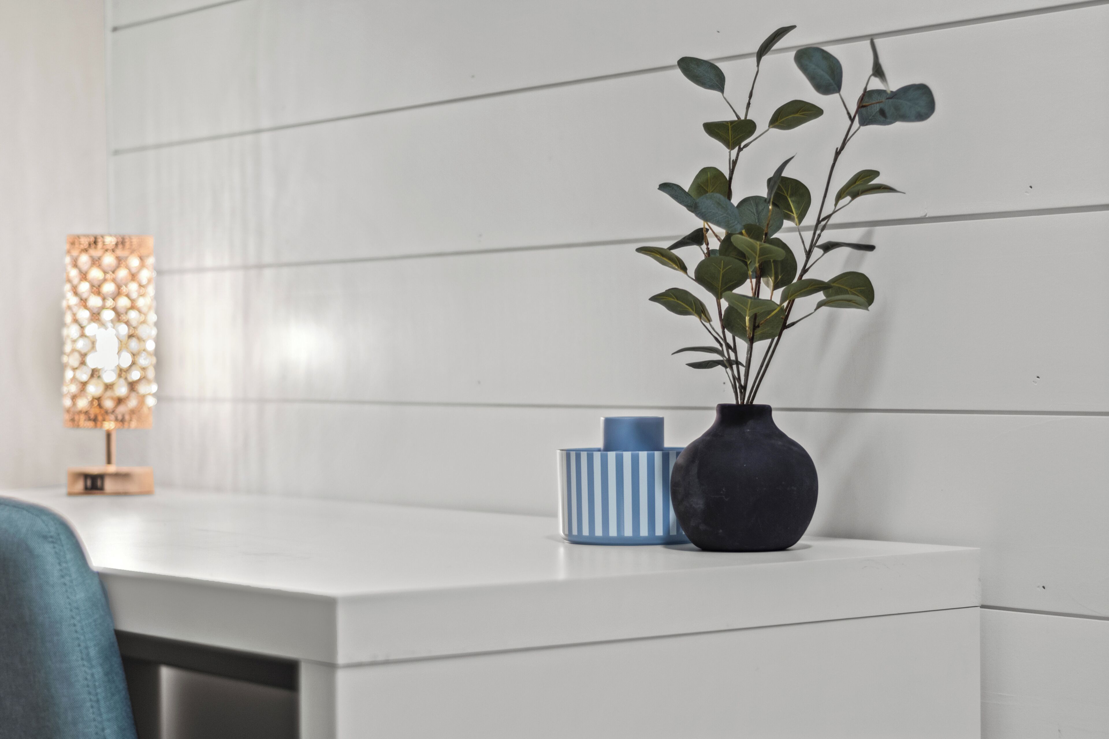 A white bedside table with a dark vase containing green leaves, a blue and white striped container, and a decorative lamp with a patterned shade.