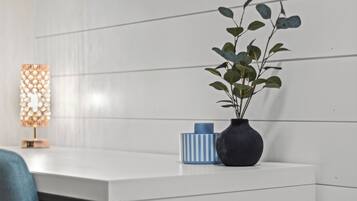 A white bedside table with a dark vase containing green leaves, a blue and white striped container, and a decorative lamp with a patterned shade.
