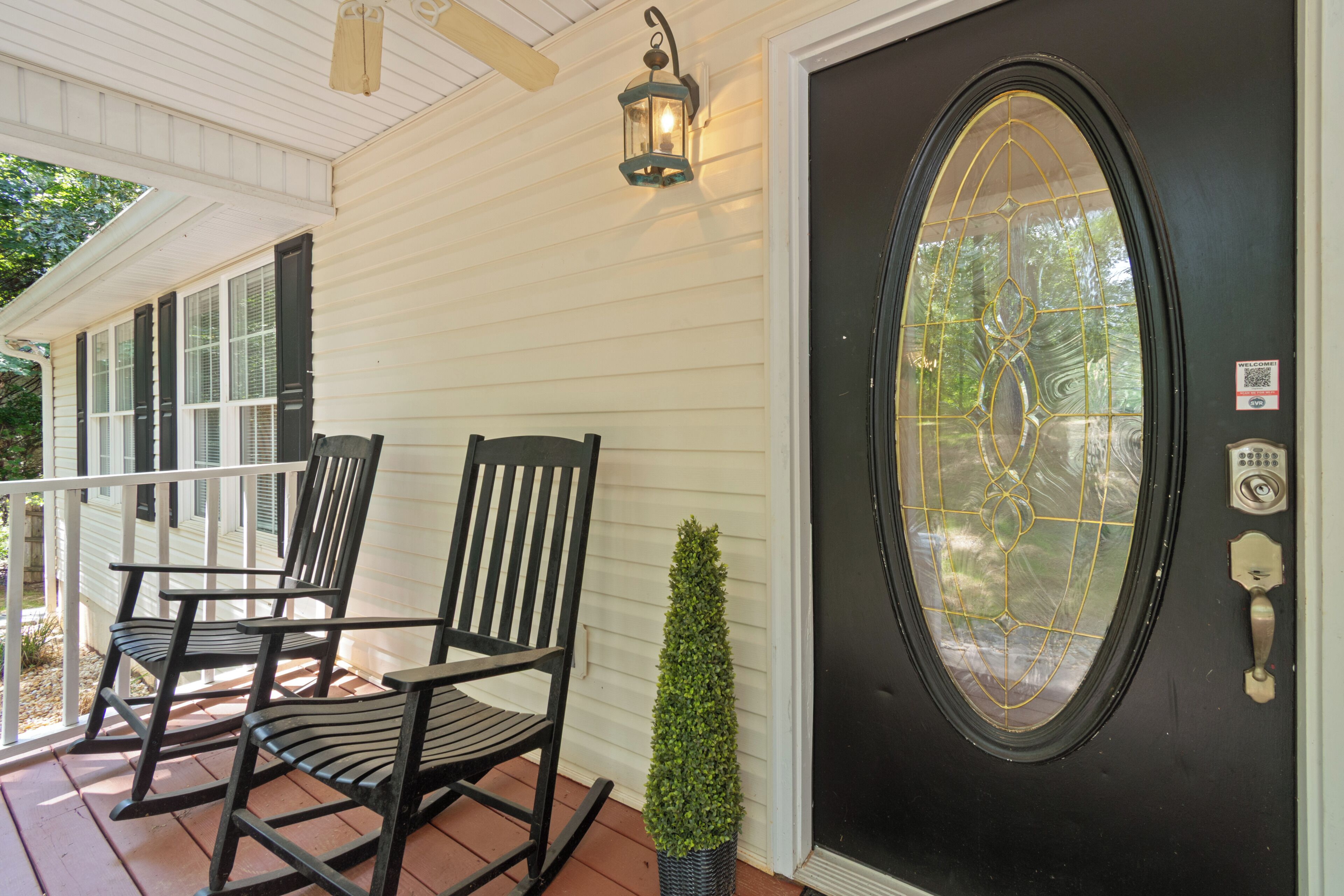 A porch with two rocking chairs, a small potted plant, and a black door with a decorative glass window.