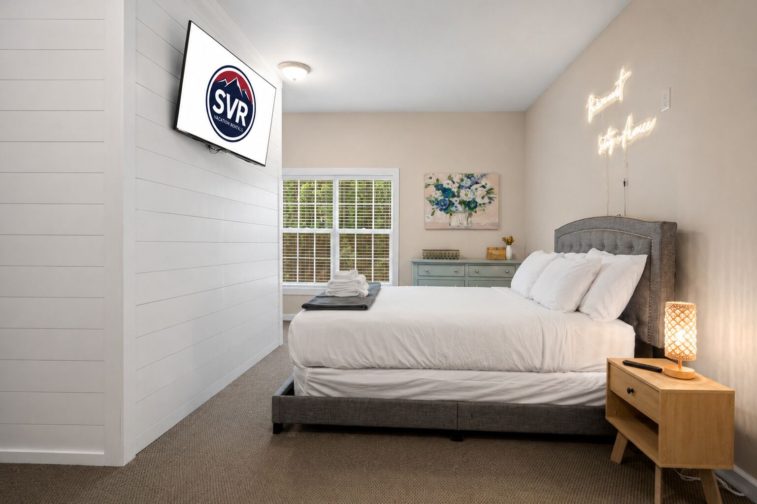 A bedroom with a bed, bedside table, nightstand, wall-mounted TV, window with blinds, and a painting.