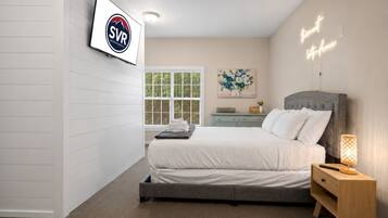 A bedroom with a bed, bedside table, nightstand, wall-mounted TV, window with blinds, and a painting.