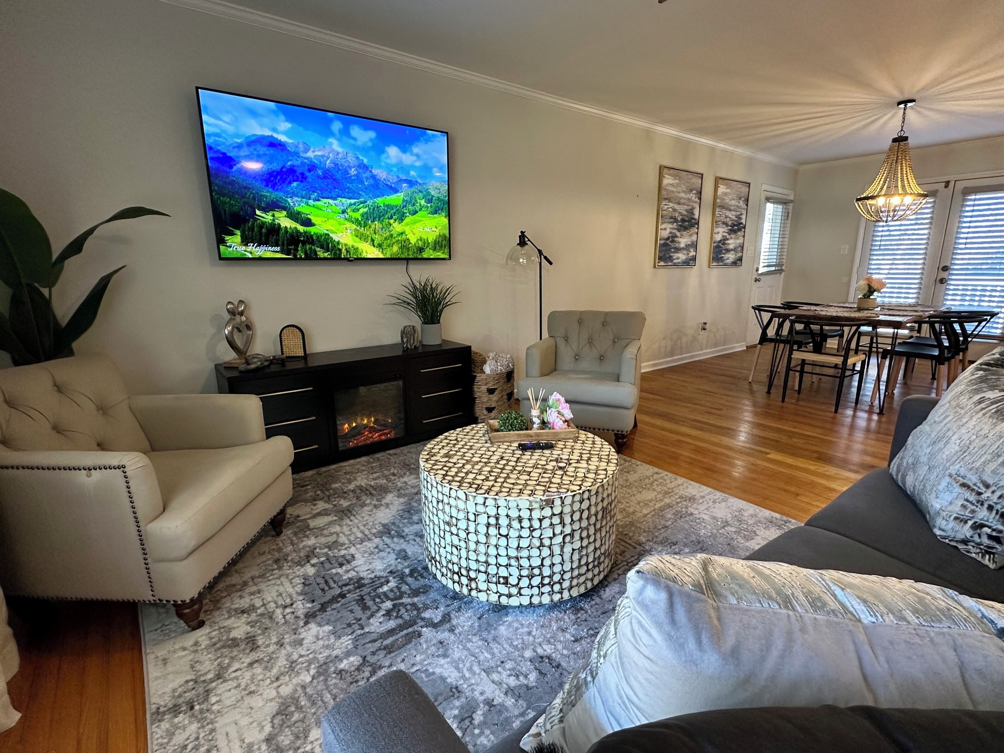 Een woonkamer met een flatscreen-televisie, een open haard, een eethoek met een tafel en stoelen, en een groot raam met zonwering.