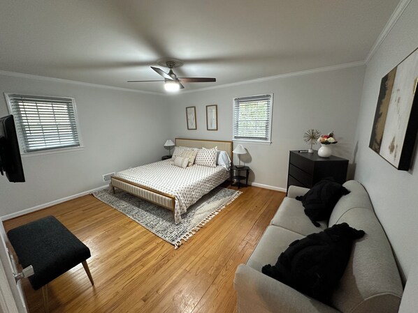 A bedroom with a bed, a ceiling fan, a nightstand, a lamp, a dresser, a window, and a wall-mounted TV.