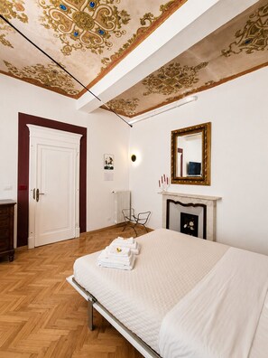 A bedroom with a bed, a dresser, a chair, a fireplace, and a ceiling with ornate designs. - Il Salotto Suite (Bologna)