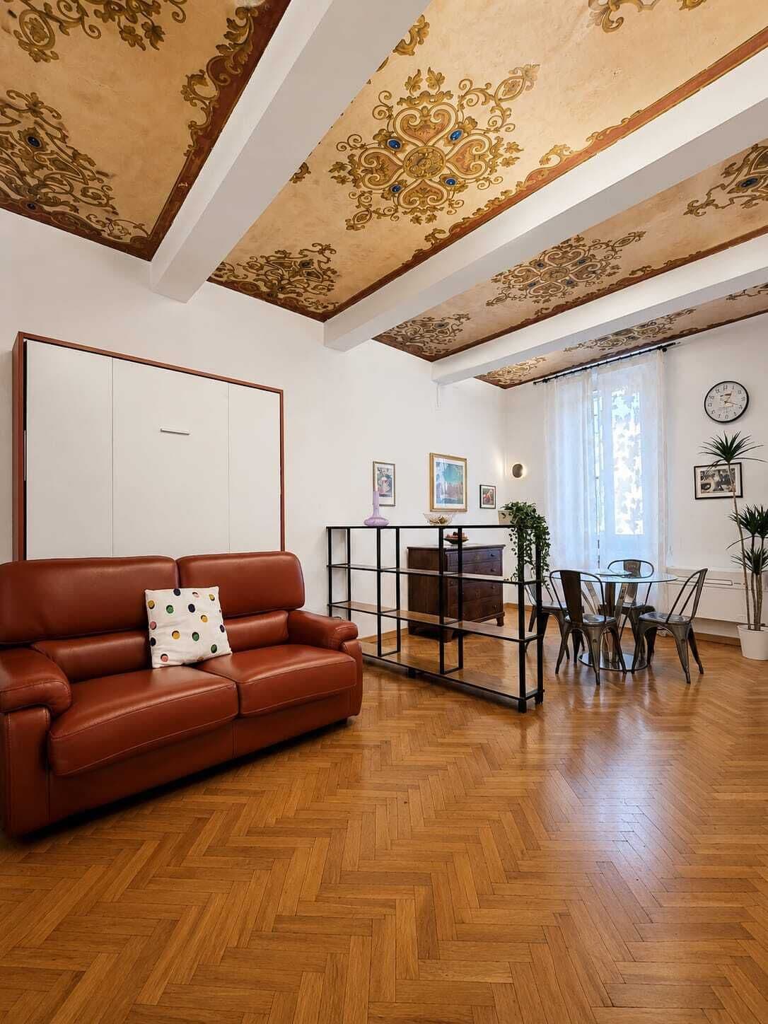 A spacious living room with a herringbone wood floor, a brown leather sofa with a polka-dotted pillow, a glass coffee table, and a dining area with metal chairs and a glass table.