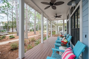 A covered porch with blue chairs and a ceiling fan, overlooking a landscaped area with trees and a path. - Chic WaterColor Getaway with 4 Suites Near Seaside and 30A. Bikes and Wraparound (Santa Rosa Beach)