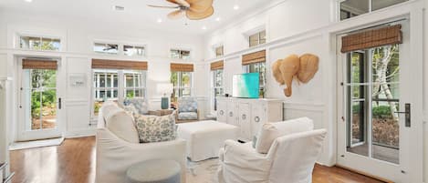 A spacious living area with a ceiling fan, white furniture, and large windows.