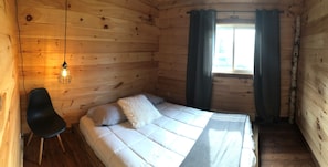 A wooden cabin room with a bed, a chair, a window with curtains, and a hanging light fixture.