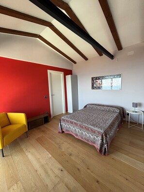 A bedroom with a bed, a yellow chair, a red wall, and wooden beams on the ceiling. - Stunning Lake View, Peaceful Green Hillside Setting, 10 Minutes from the Beaches (Toscolano Maderno)