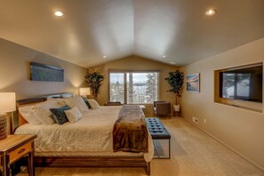 Une chambre à coucher avec un grand lit, un banc, un bureau et une fenêtre donnant sur des arbres.