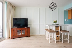 Een kamer met een televisie op een houten kast, een witte tafel met krukken en een hangende lamp.