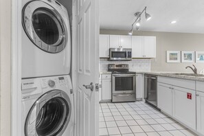 Un cuarto de lavado con lavadora y secadora de carga frontal, microondas, horno y lavavajillas en un área de cocina con gabinetes blancos y piso de baldosas.