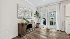 A spacious room with hardwood flooring, a wooden console table with decorative items, a large mirror, and a view of a blue house through a sliding glass door. - Luxury Furnished 3BR Townhome-Walk to Rio and Crown Plaza Garage-Modern Finishes (Gaithersburg)