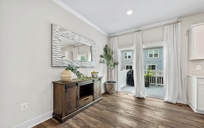 A spacious room with hardwood flooring, a wooden console table with decorative items, a large mirror, and a view of a blue house through a sliding glass door. - Luxury Furnished 3BR Townhome-Walk to Rio and Crown Plaza Garage-Modern Finishes (Gaithersburg)