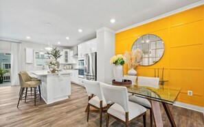 A modern kitchen with white cabinetry, a central island, and a dining area with a glass table and chairs. - Luxury Furnished 3BR Townhome-Walk to Rio and Crown Plaza Garage-Modern Finishes (Gaithersburg)