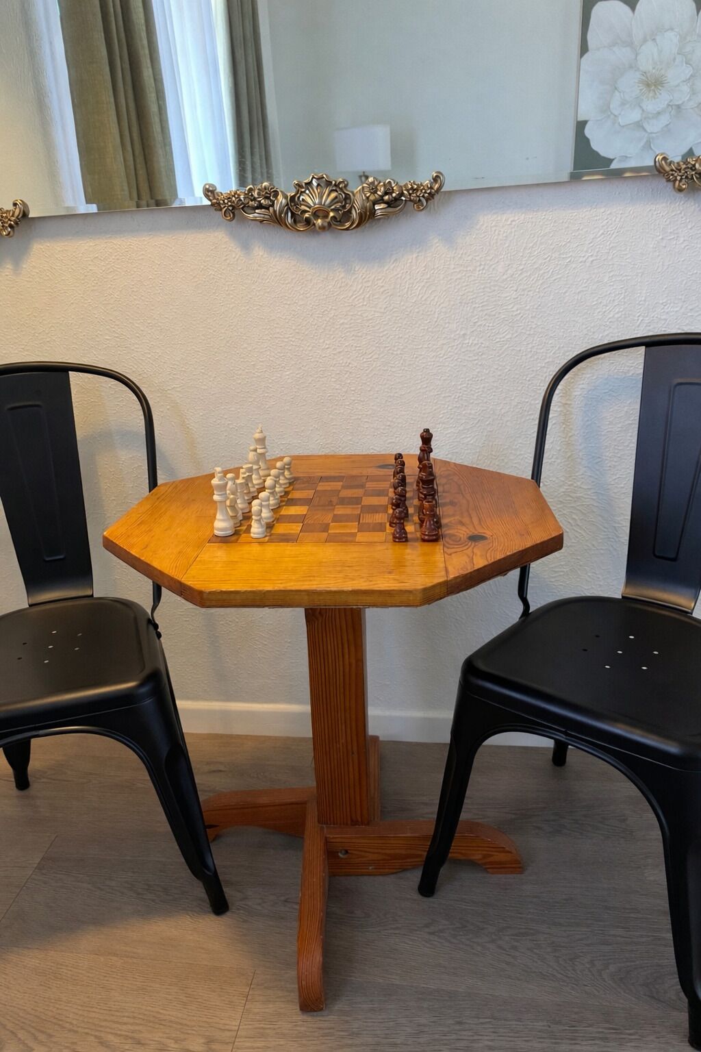 A chessboard set on a wooden table with two black chairs.