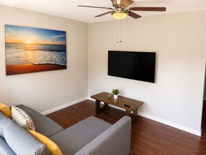 Un salon comprenant un canapé, un téléviseur à écran plat, un ventilateur de plafond et un tableau représentant un coucher de soleil sur l’océan.