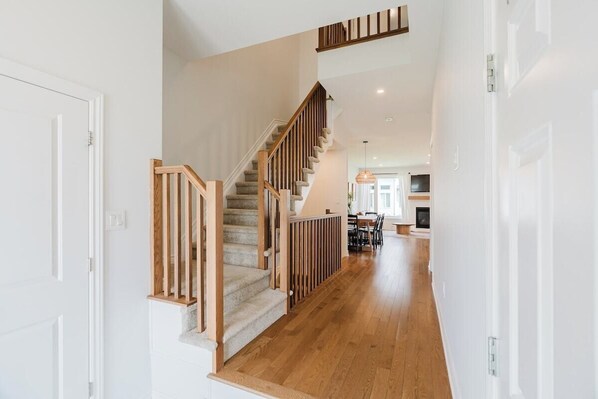 A staircase with wooden railings leading to a second floor, a dining area with chairs and a table, and a kitchen area with a TV.
