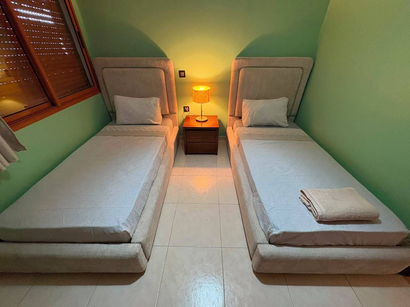 A twin room with two single beds, a nightstand with a lamp, and a window with blinds.