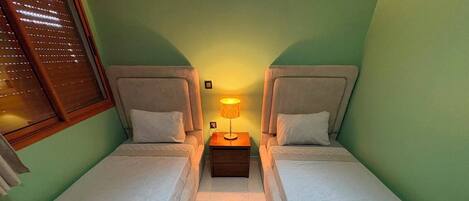 A twin room with two single beds, a nightstand with a lamp, and a window with blinds.