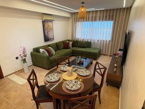 Una sala de estar con un sofá modular, una mesa de comedor con individuales y un televisor sobre un mueble.