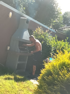Una persona usando un horno grande al aire libre, rodeada de follaje y una estructura de edificio.