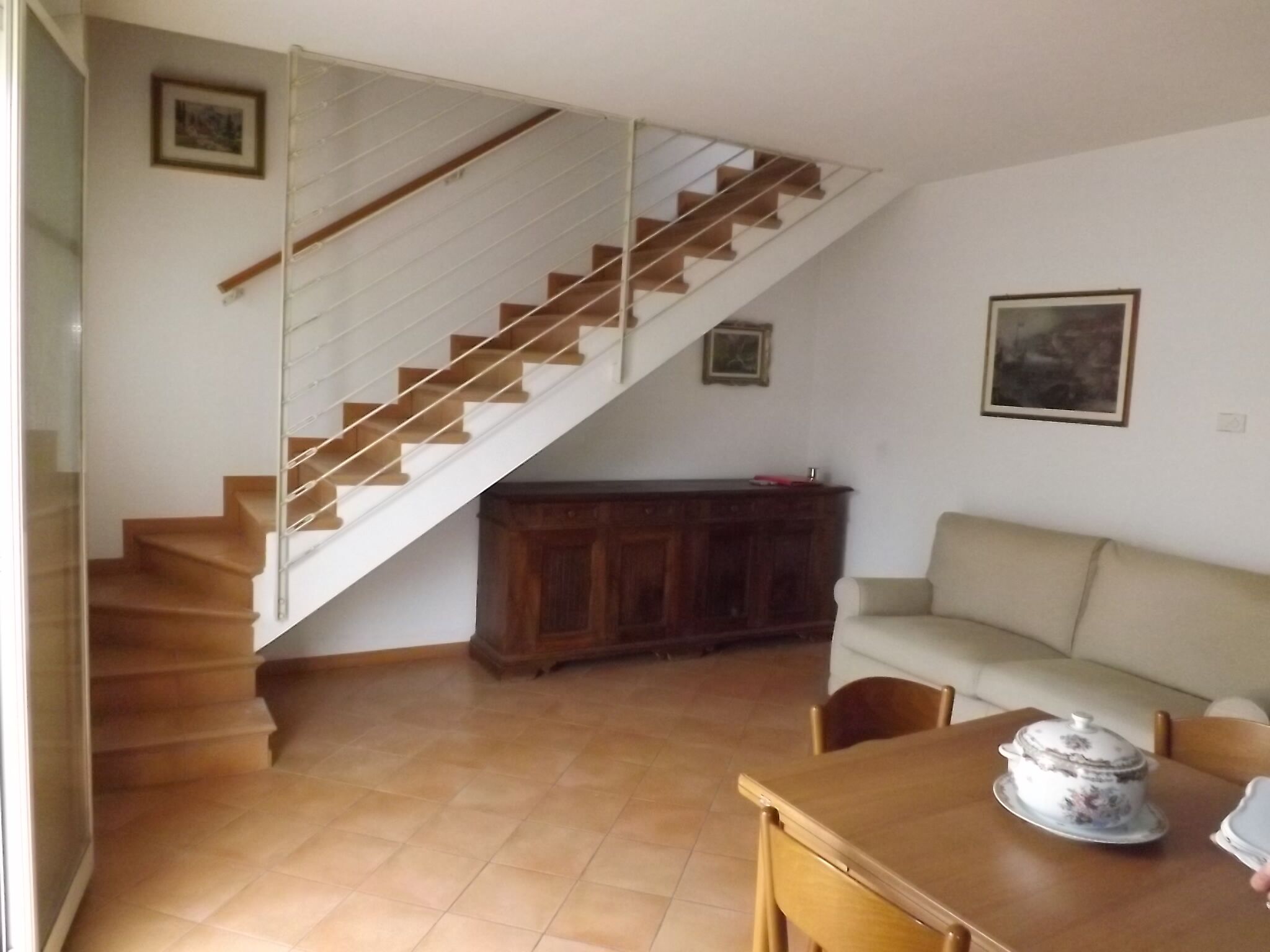 Un salon avec un escalier, une armoire en bois, un canapé beige et une table à manger sur laquelle se trouve une casserole couverte.