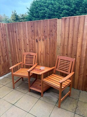 Two wooden chairs with slatted backs and armrests, placed on a tiled patio with a wooden fence in the background.
