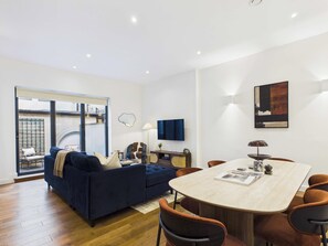 A modern living room with a dark blue sofa, a round dining table, and a large window. - Spacious Baker Street Apartment with Balcony (Greater London)