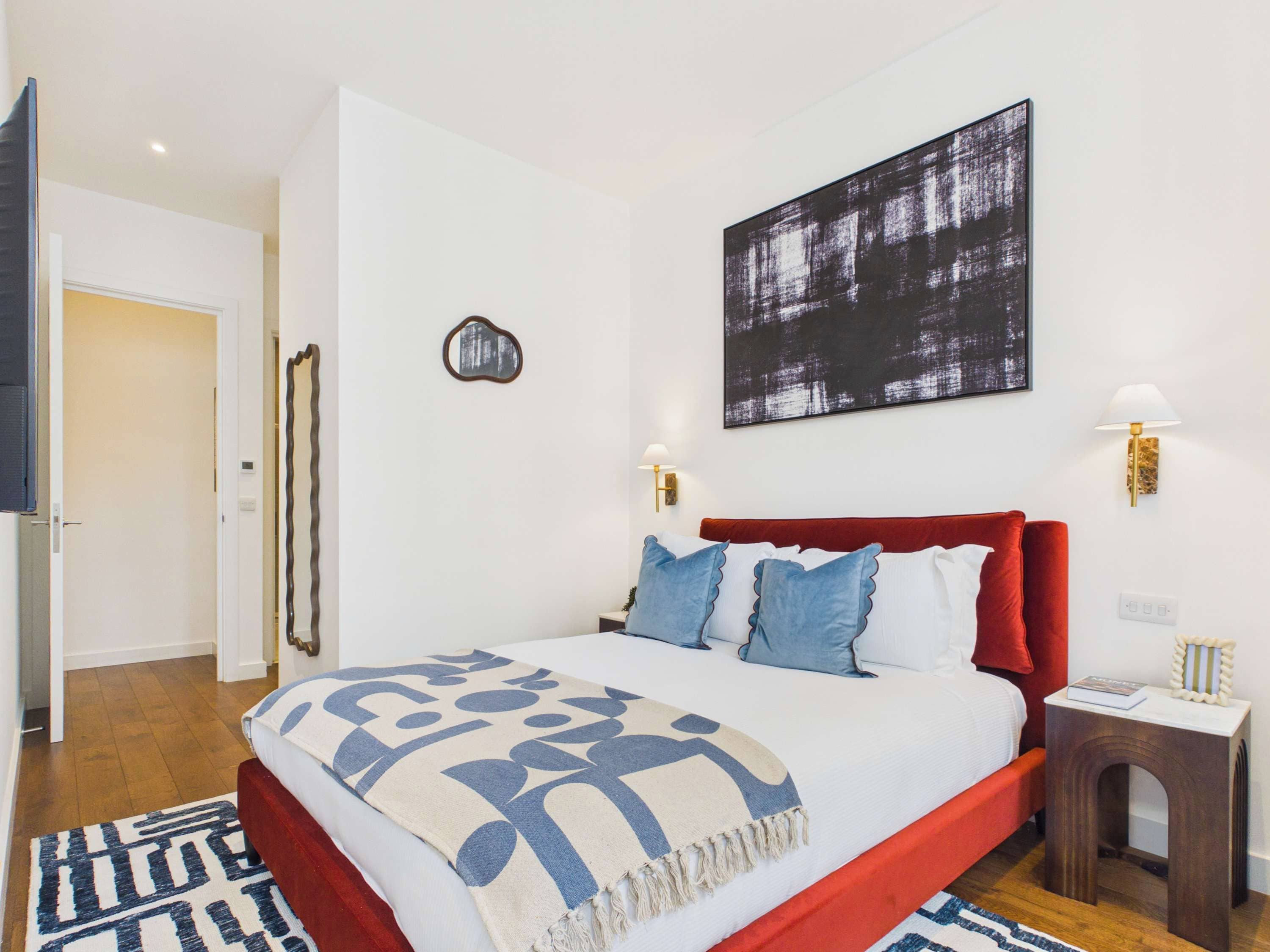 Quarto moderno com cabeceira vermelha, uma grande pintura abstrata, duas luminárias de parede e um tapete estampado.
