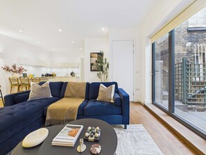 A modern living room with a sectional sofa, a coffee table with decorative items, and a view of a cityscape through large windows. - Spacious Baker Street Apartment with Balcony (Greater London)