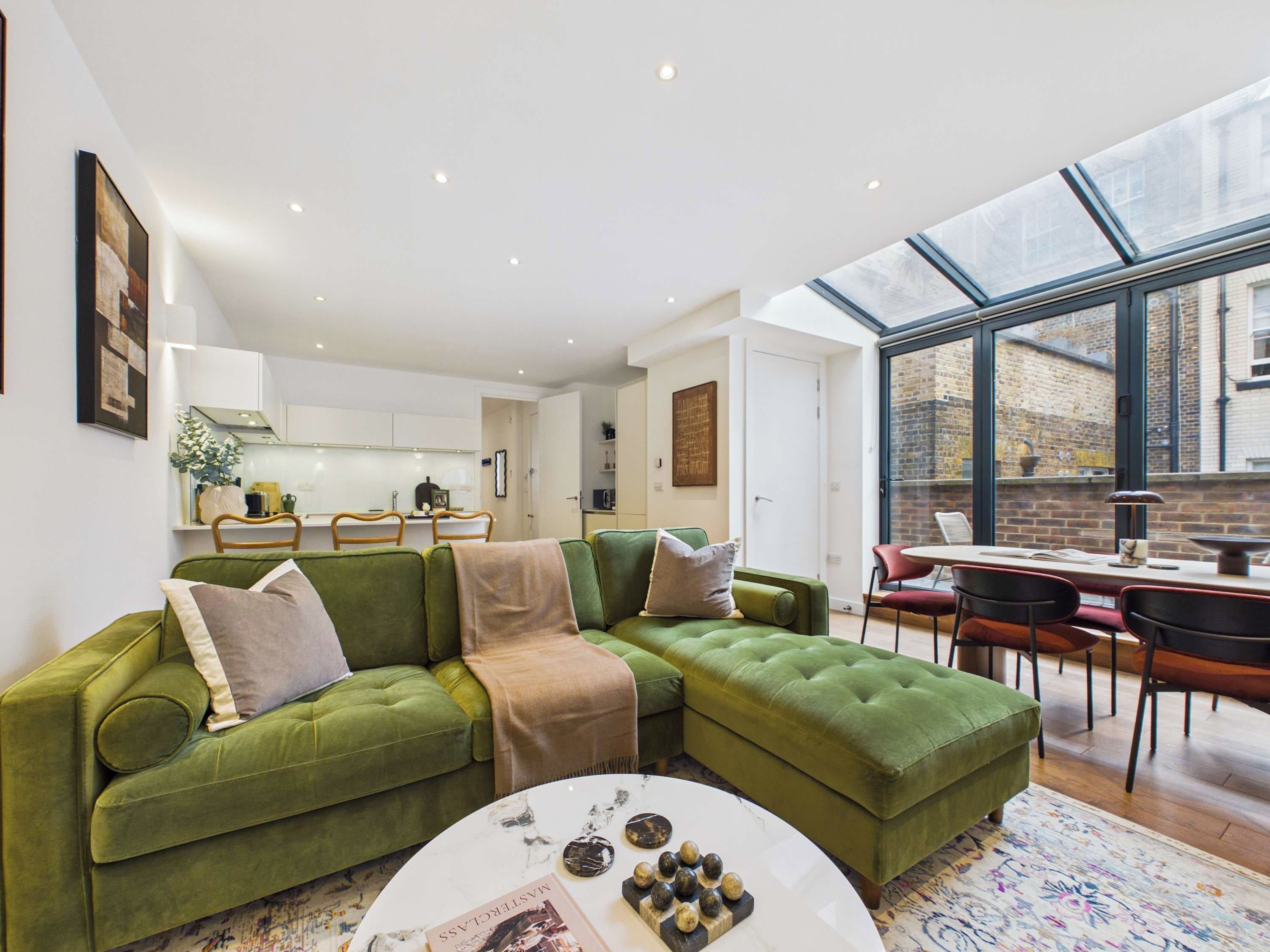 A modern living room with a green sectional sofa, a dining area with a table and chairs, and a kitchen in the background.