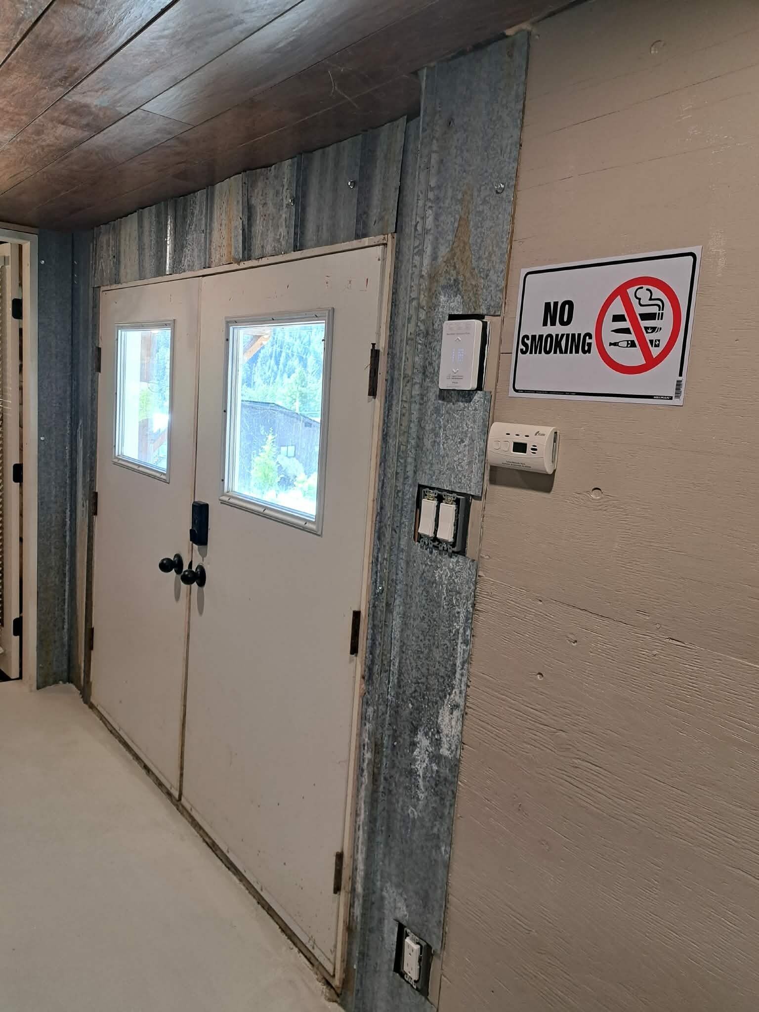 Un pasillo con dos puertas, un cartel de prohibido fumar y un detector de humo.