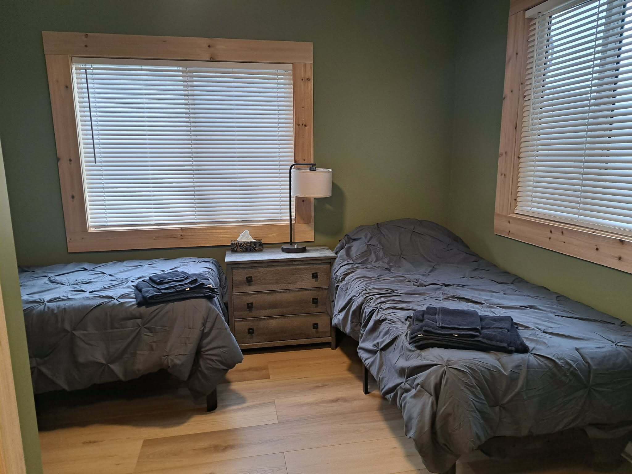 Una habitación pequeña y acogedora con dos camas individuales, una mesita de noche de madera, una lámpara y una ventana con persianas.