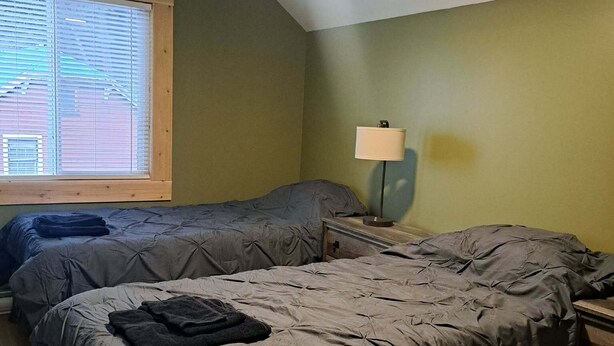 A bedroom with two beds, a ceiling fan, a window with blinds, and a bedside table with a lamp.