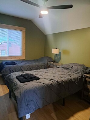 A bedroom with two beds, a ceiling fan, a window with blinds, and a bedside table with a lamp.